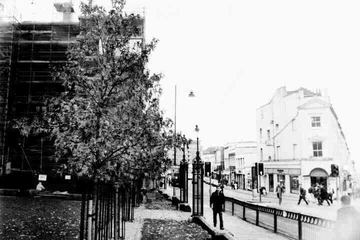 A black and white image of a street in Bristol. A person is walking towards the camera, while behind people are crossing the road.