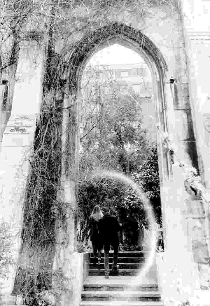 A black and white photo of two people walking through an arch. They are framed by a white semi-circle caused by a kink in the film.