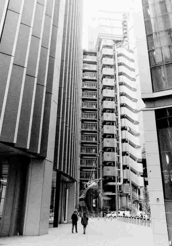 A black and white photo of two people walking through the streets, surrounded on all sides by towering office buildings.
