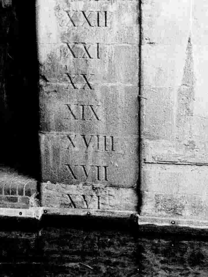 A black and white photo of Roman numerals from 16 to 22 carved into a wall under a bridge.