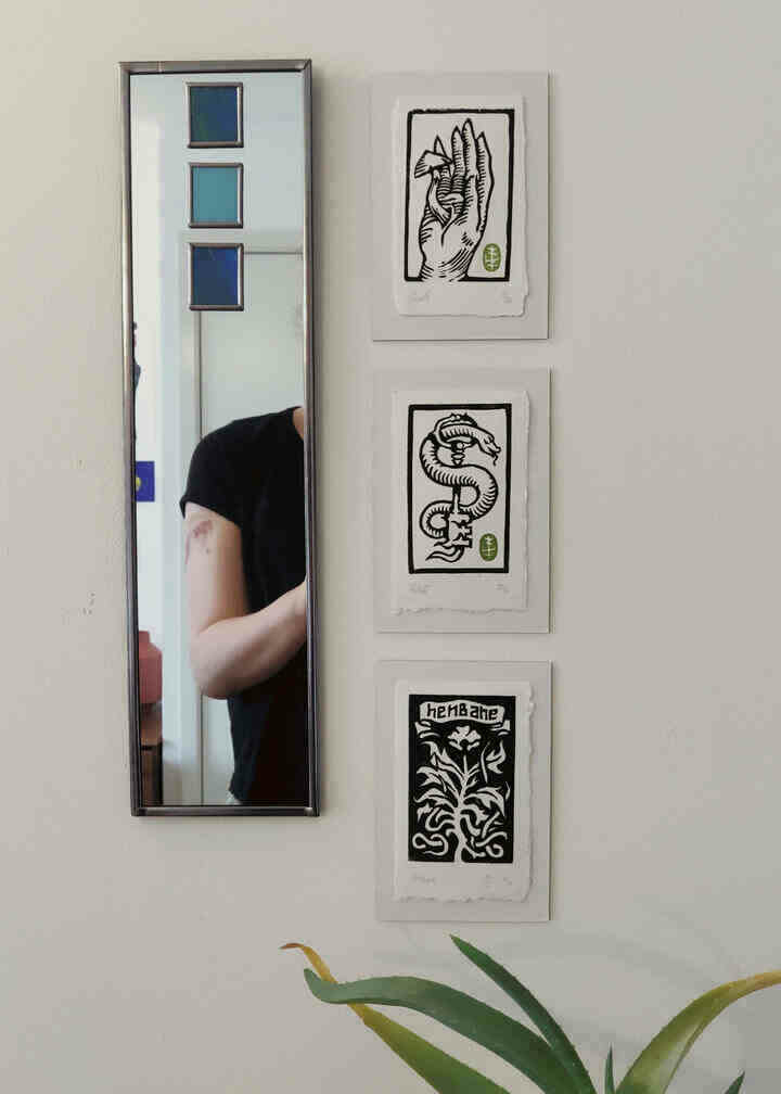 Three linocut prints by Ritual Dust displayed next to a mirror. The top one is a mushroom growing out of a hand. The middle is a snake and a key, representing the goddess Hekate. The bottom is of the plant henbane.